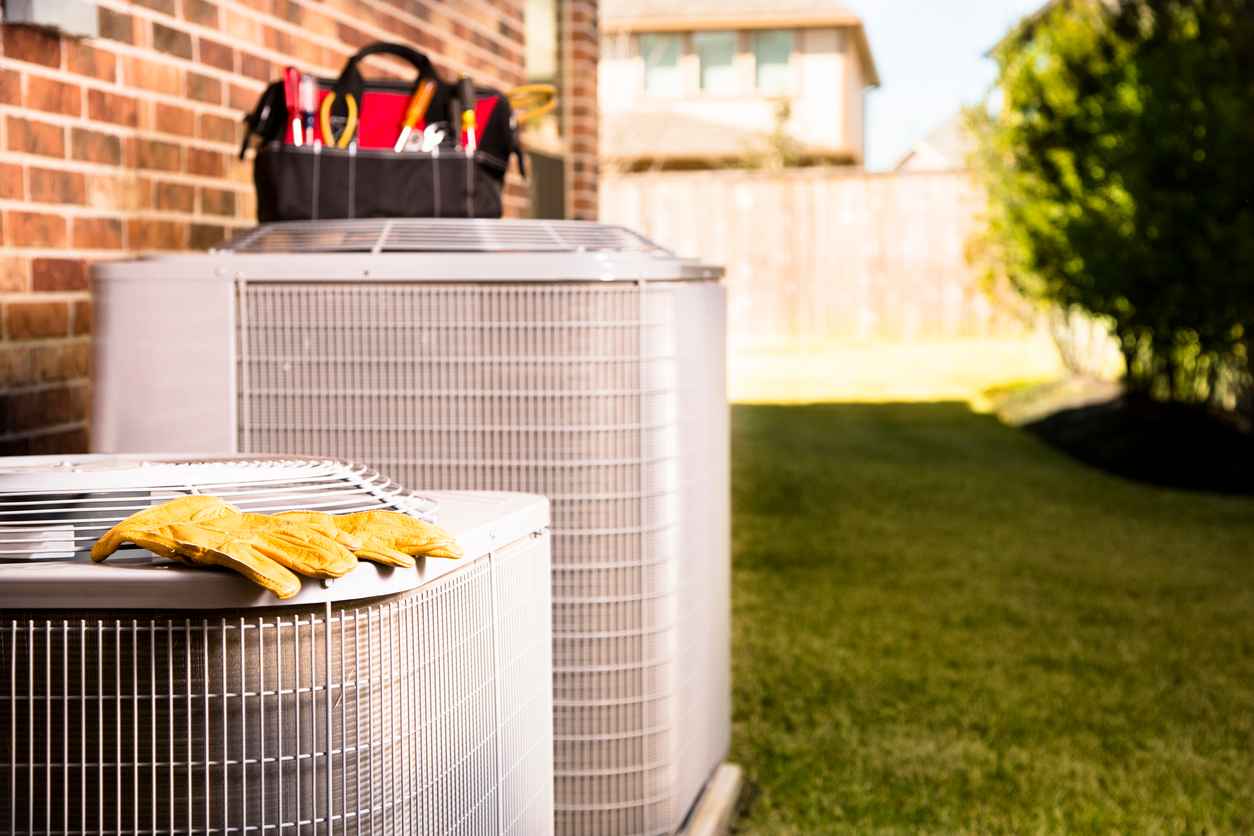 Bag of repairman's work tools, gloves on top of air conditioner units outside a brick home.